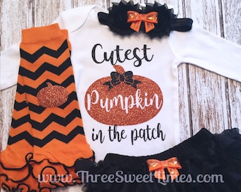 Może przedstawiać: Biały śpioszek z napisem "Cutest Pumpkin in the patch" i błyszczącą pomarańczową grafiką dyni. Śpioszek jest połączony z czarną tiulową spódniczką, pomarańczowo-czarnymi legginsami w chevron i czarną opaską z pomarańczową kokardką.