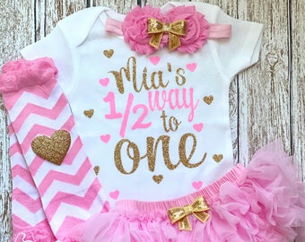 Può includere: Un completo per neonata rosa e oro con un body bianco che dice "Mia's 1/2 way to one" in glitter dorati. Il completo include anche un tutù rosa, scaldagambe a righe rosa e bianche e una fascia rosa con un fiocco dorato.