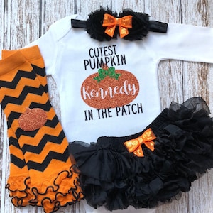 May include: A white onesie with the text "Cutest Pumpkin Kennedy in the Patch" and a glittery orange pumpkin graphic. The onesie is paired with a black tulle tutu with an orange bow, black and orange chevron leg warmers, and a black headband with an orange bow.