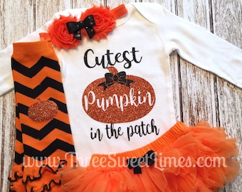 Może przedstawiać: Pomarańczowy i czarny strój dla niemowlęcia z białą koszulką z długim rękawem, na której napisano "Cutest Pumpkin in the patch" z grafiką błyszczącej dyni. Strój zawiera również tiulową spódniczkę, legginsy i opaskę na głowę.