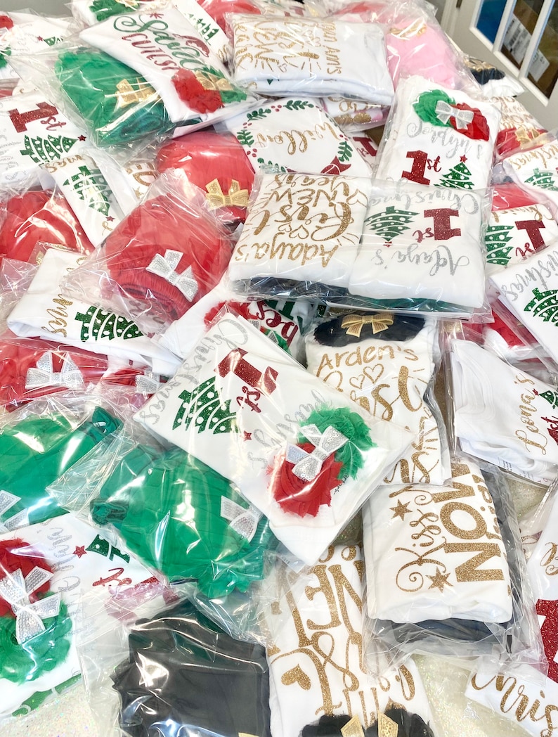 Puede incluir: Una pila de bodies blancos con varios dise&ntilde;os navide&ntilde;os y detalles de purpurina. Los bodies est&aacute;n empaquetados en bolsas de pl&aacute;stico transparentes. Algunos de los dise&ntilde;os incluyen "Arden's 1st Christmas", "Adaya's First New Year", "Kiss My  Mistletoe", y "Jerdyn's 1st Christmas".