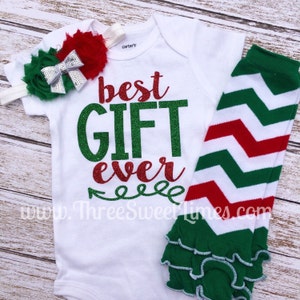 Puede incluir: Un body blanco para beb&eacute; con el texto "best GIFT ever" en purpurina verde y roja. El body se combina con un calienta piernas de chevron verde y rojo con un volante blanco en la parte inferior y una diadema de flores verde y plata.