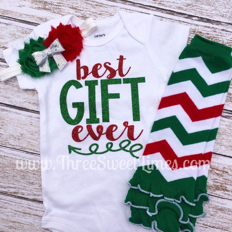 Puede incluir: Un body blanco para beb&eacute; con el texto "best GIFT ever" en purpurina verde y roja. El body se combina con un calienta piernas de chevron verde y rojo con un volante blanco en la parte inferior y una diadema de flores verde y plata.