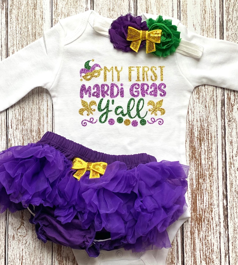 May include: A baby outfit set for Mardi Gras. The set includes a white bodysuit with the text "My First Mardi Gras Y'all" in gold, purple, and green glitter, a purple tulle tutu with a gold bow, and a headband with a purple and green flower and a gold bow.