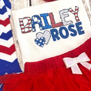 May include: A white bodysuit with the name "BAILEY ROSE" in red, white, and blue glitter letters with a red, white, and blue heart design. The bodysuit is paired with a red tulle skirt with a white sequin bow and a red and white striped headband with a white sequin bow.