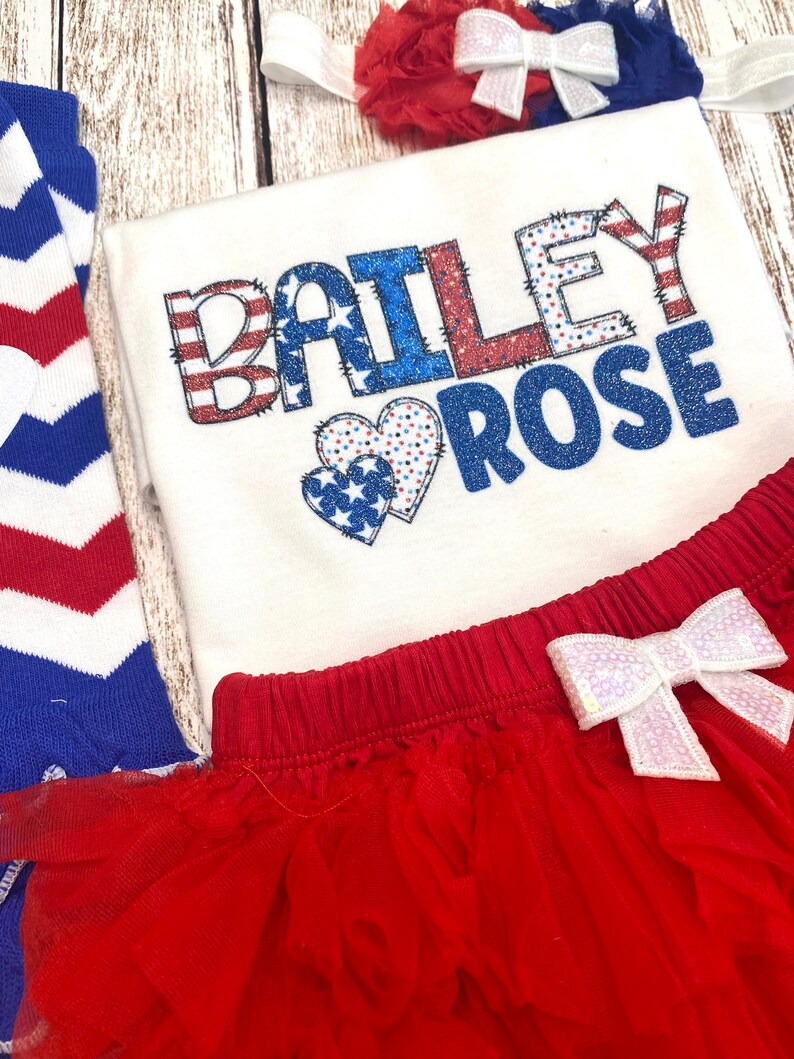 May include: A white bodysuit with the name "BAILEY ROSE" in red, white, and blue glitter letters with a red, white, and blue heart design. The bodysuit is paired with a red tulle skirt with a white sequin bow and a red and white striped headband with a white sequin bow.