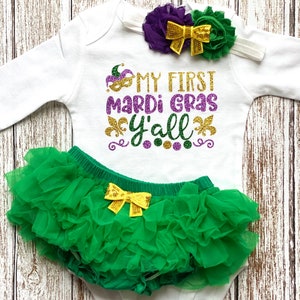 May include: A white baby onesie with the text "My First Mardi Gras Y'all" in gold glitter lettering. The onesie is paired with a green tulle tutu and a purple and green flower headband with a gold bow.