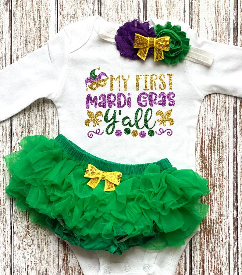 May include: A white baby onesie with the text "My First Mardi Gras Y'all" in gold glitter lettering. The onesie is paired with a green tulle tutu and a purple and green flower headband with a gold bow.