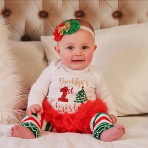 Puede incluir: Una ni&ntilde;a peque&ntilde;a con un body blanco con el texto "Brooklyn's 1st Christmas" en purpurina dorada. Lleva una falda de tul a rayas rojas y verdes y una diadema con un lazo rojo y verde.