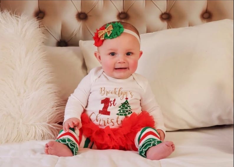 Puede incluir: Una ni&ntilde;a peque&ntilde;a con un body blanco con el texto "Brooklyn's 1st Christmas" en purpurina dorada. Lleva una falda de tul a rayas rojas y verdes y una diadema con un lazo rojo y verde.