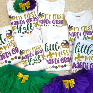 May include: Five white onesies with gold glitter text that reads "My First Mardi Gras Y'all" and "Little Miss Mardi Gras".  There are two green and purple tutus with gold bows.