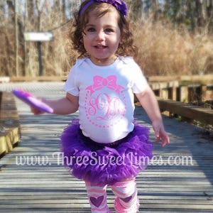 May include: A young girl wearing a white bodysuit with a pink bow and a monogrammed letter 'S' in a circle. She is also wearing a purple tulle skirt, pink and white striped leg warmers with pink hearts, and a purple headband. The girl is standing on a wooden walkway.