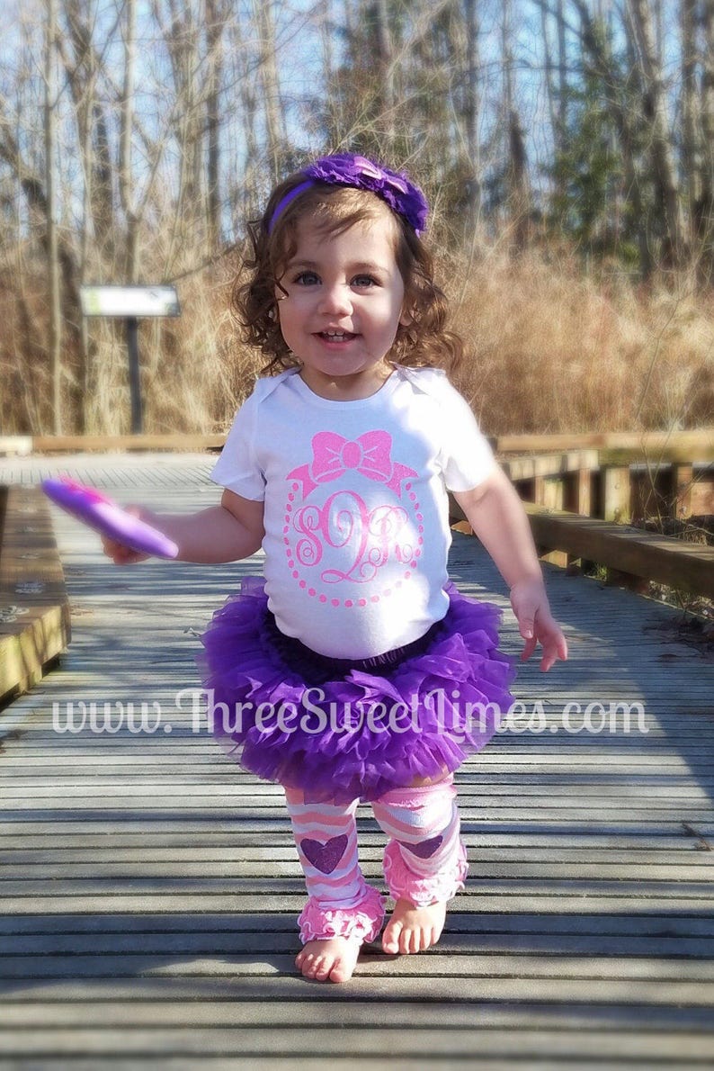 May include: A young girl wearing a white bodysuit with a pink bow and a monogrammed letter 'S' in a circle. She is also wearing a purple tulle skirt, pink and white striped leg warmers with pink hearts, and a purple headband. The girl is standing on a wooden walkway.