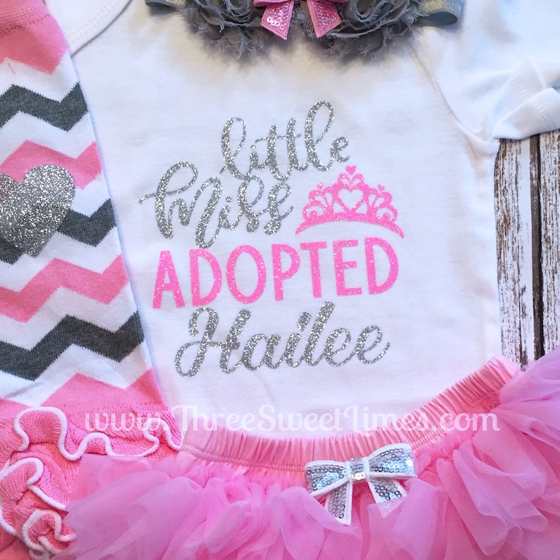 May include: A white onesie with silver glitter text that reads "little miss adopted Hailee" with a pink crown above the word "adopted". The onesie is paired with a pink tutu and gray and pink chevron leggings.