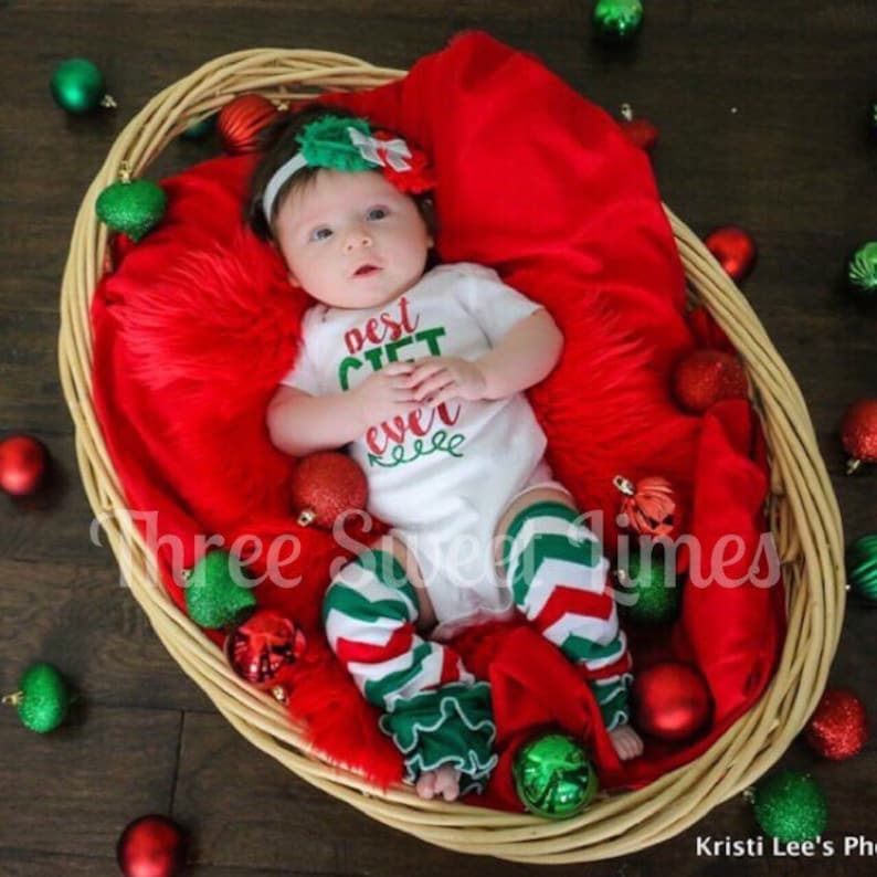 Puede incluir: Un beb&eacute; con un body blanco con el texto "Best Gift Ever" en letras verdes y rojas. El beb&eacute; lleva un cubre piernas a rayas verdes y rojas con un volante verde. El beb&eacute; est&aacute; acostado en una cesta de mimbre con una manta roja y rodeado de adornos navide&ntilde;os rojos y verdes.
