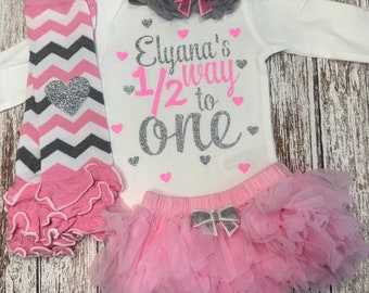 May include: A baby girl's outfit featuring a white long-sleeve bodysuit with the text "Elyana's 1/2 way to One" in pink and silver glitter. The set includes a pink tutu skirt, a gray headband with a bow, and pink and gray leg warmers.