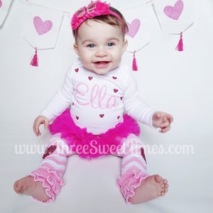 May include: A baby girl wearing a white bodysuit with pink glitter hearts and the name "Ella" on the front. She is also wearing a pink tulle skirt and pink and white striped leggings with pink ruffles at the bottom. The baby is sitting on a white background with pink heart-shaped decorations.