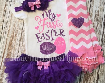 May include: A white onesie with the text "My First Easter" in purple glitter, a pink glitter egg with the year 2017, and a purple glitter circle with the name "Addyson". The onesie is paired with a purple tulle skirt with a pink bow, a purple headband with a pink bow, and pink and white chevron leg warmers with a purple glitter heart.