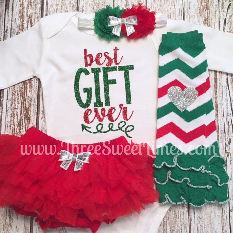 Puede incluir: Un conjunto de ropa de beb&eacute; de Navidad con un body blanco que dice "best GIFT ever" en purpurina verde y roja, un tut&uacute; rojo de tul con un lazo plateado, polainas a rayas verdes y blancas con un coraz&oacute;n plateado y una diadema de flores verde y roja con un lazo plateado.