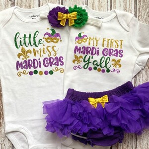 May include: Two white baby onesies with purple, gold, and green glitter text. One onesie says "little miss mardi gras" and the other says "my first mardi gras y'all". A purple tulle tutu with a gold bow is included.