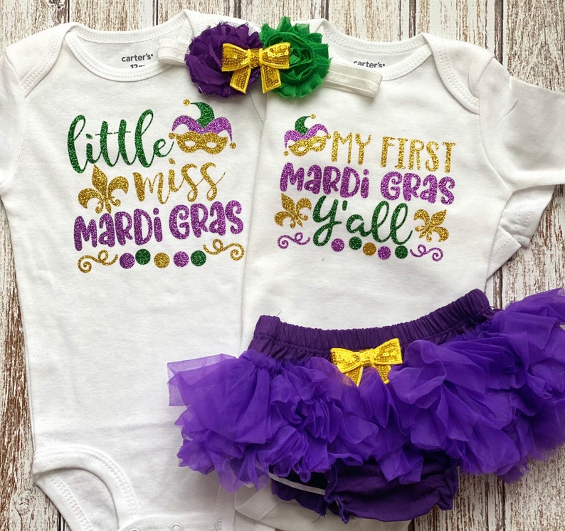 May include: Two white baby onesies with purple, gold, and green glitter text. One onesie says "little miss mardi gras" and the other says "my first mardi gras y'all". A purple tulle tutu with a gold bow is included.