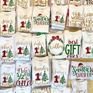 Puede incluir: Una colecci&oacute;n de bodies blancos con dise&ntilde;os de purpurina para una primera Navidad. Los bodies presentan varios dise&ntilde;os, incluyendo un &aacute;rbol de Navidad, Santa Claus y las palabras "primera Navidad".
