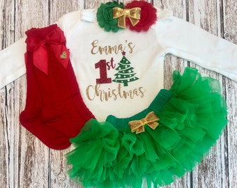 Puede incluir: Un mono blanco de manga larga con el texto "Emma's 1st Christmas" en letras doradas de purpurina y un gráfico de árbol de Navidad verde. El mono se combina con una falda de tul verde con un lazo dorado, leotardos de punto rojo con un lazo rojo y una diadema de flores roja y verde con un lazo dorado.