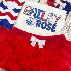 May include: A red, white, and blue baby outfit with a white onesie that says "BAILEY ROSE" in red, white, and blue letters with two red, white, and blue hearts. The outfit also includes a red tulle tutu with a white bow, a red and white striped leg warmer, and a red headband with a white bow.