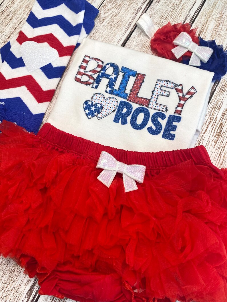 May include: A red, white, and blue baby outfit with a white onesie that says "BAILEY ROSE" in red, white, and blue letters with two red, white, and blue hearts. The outfit also includes a red tulle tutu with a white bow, a red and white striped leg warmer, and a red headband with a white bow.