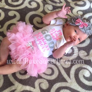 May include: A baby girl wearing a white onesie with the text "I'm too pretty that God answers prayers" in pink and silver glitter. She is also wearing a pink tulle tutu and a gray headband with a pink bow.