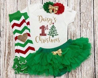 Puede incluir: Un body blanco con el texto "Daisy's 1st Christmas" en letras doradas con purpurina y un árbol de Navidad verde con purpurina. El body se combina con un tutú de tul verde con un lazo dorado, polainas de chevron verde y rojo con un corazón dorado y una diadema con lazo rojo y dorado.