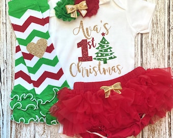 Puede incluir: Un body blanco con el texto "Ava's 1st Christmas" en letras doradas con purpurina y un gráfico de árbol de Navidad verde. El body se combina con un tutú rojo de tul, calentadores de piernas de chevron verde y rojo y una diadema de flores verde y dorada.
