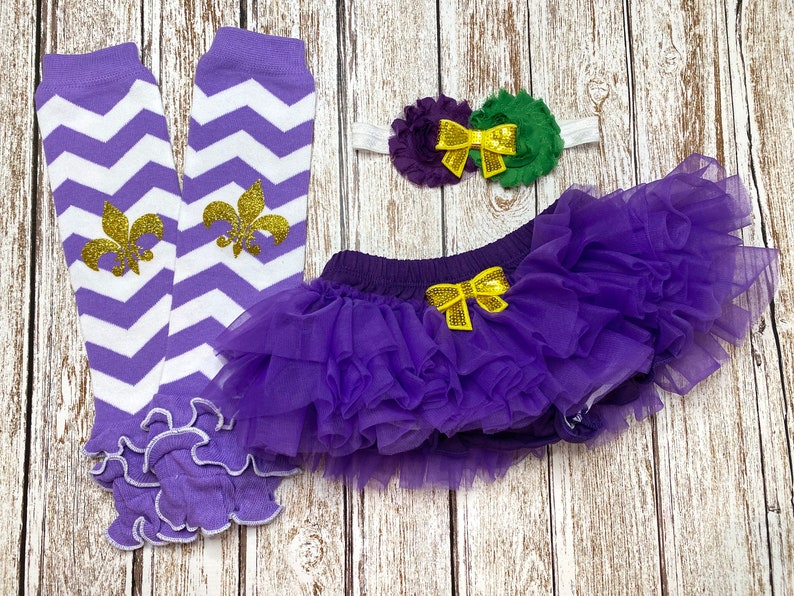 May include: Purple and white chevron leg warmers with gold fleur-de-lis accents, a purple tulle tutu with a yellow bow, and a purple and green flower headband with a yellow bow.