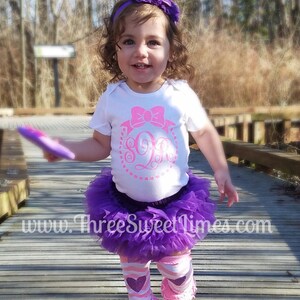 May include: A baby girl wearing a white shirt with a pink bow and a monogram, a purple tulle tutu, and pink and purple striped leg warmers with pink ruffles. The shirt has a pink glitter design with the letters "SO" and a pink bow.