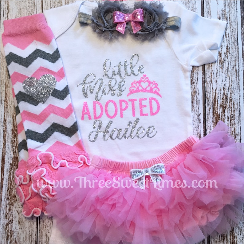 May include: A pink and gray chevron leg warmer with a silver heart, a white onesie with the text "little miss adopted Hailee" in pink and silver glitter, a pink tulle skirt with a silver bow, and a gray flower headband with a pink bow.