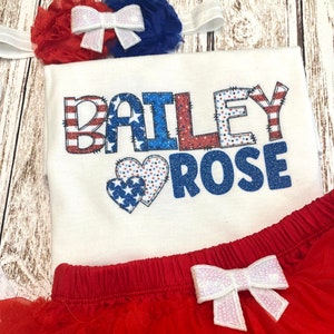 May include: A white onesie with the name "BAILEY ROSE" in red, white, and blue glitter letters with a red, white, and blue heart design. The onesie is paired with a red tulle skirt with a white bow.