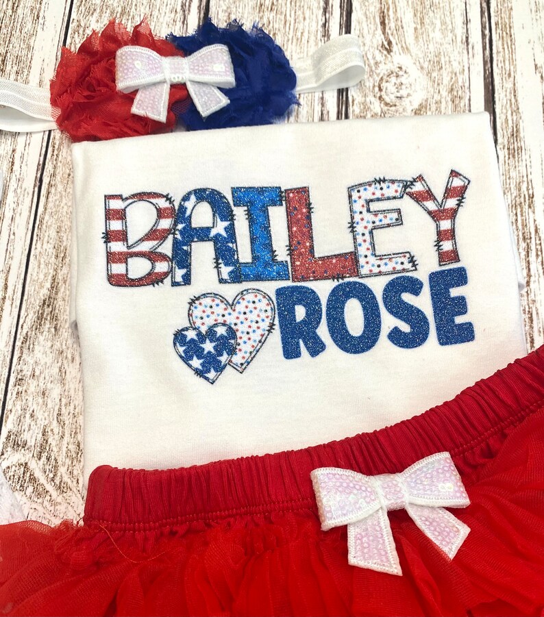 May include: A white onesie with the name "BAILEY ROSE" in red, white, and blue glitter letters with a red, white, and blue heart design. The onesie is paired with a red tulle skirt with a white bow.