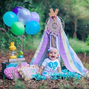 May include: A baby girl wearing a blue and white shirt with the text "1/2 Alissa" sits inside a teepee decorated with a dreamcatcher and colorful balloons.