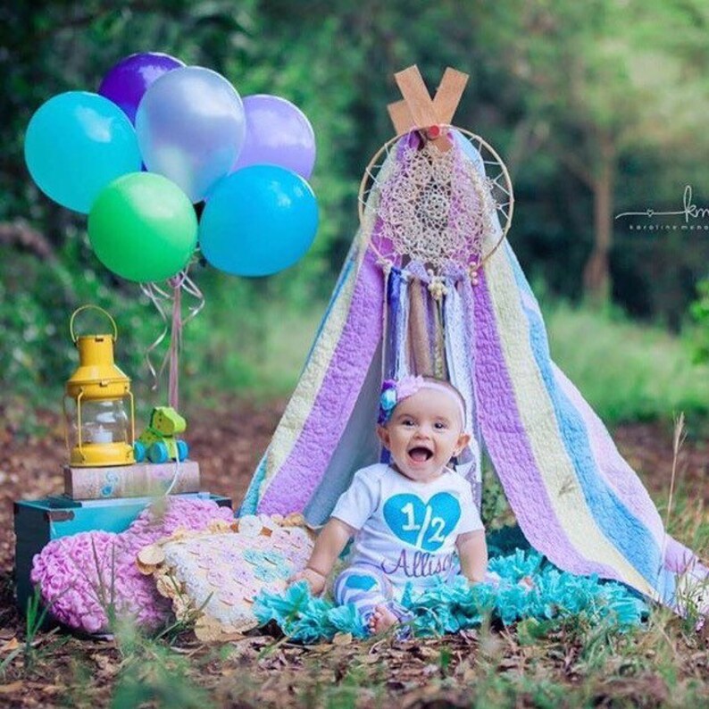 May include: A baby girl wearing a blue and white shirt with the text "1/2 Alissa" sits inside a teepee decorated with a dreamcatcher and colorful balloons.