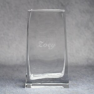 May include: A clear, rectangular glass vase with a slightly curved design. The word "Zoey" is etched into the glass. The vase is empty and sits on a gray surface.