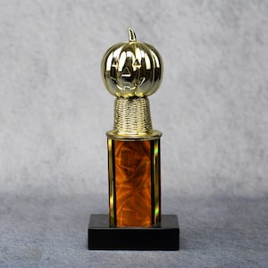 May include: A gold-colored trophy with a pumpkin topper. The trophy has a gold stem with a decorative woven pattern, a rectangular orange and black base, and a black square base. A festive award for Halloween.
