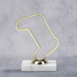 May include: A gold-colored trophy with a unique, abstract design, mounted on a white marble base. The trophy's shape is a stylized outline. The base is rectangular and the trophy is set against a neutral background.