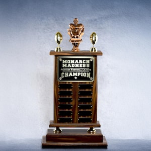 May include: A wooden trophy with a gold plaque that reads "Monarch Madness Fantasy Football League Champion". The trophy has a football player figurine on top and two footballs on the sides.