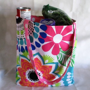 May include: A colorful tote bag with a floral pattern in pink, orange, blue, and green. The bag has a pink polka-dot strap and is filled with groceries, including a can and a bag of produce.