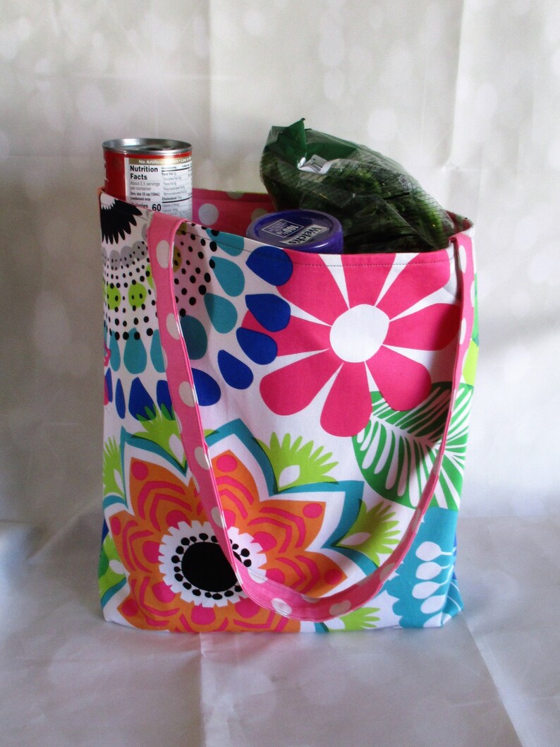 May include: A colorful tote bag with a floral pattern in pink, orange, blue, and green. The bag has a pink polka-dot strap and is filled with groceries, including a can and a bag of produce.