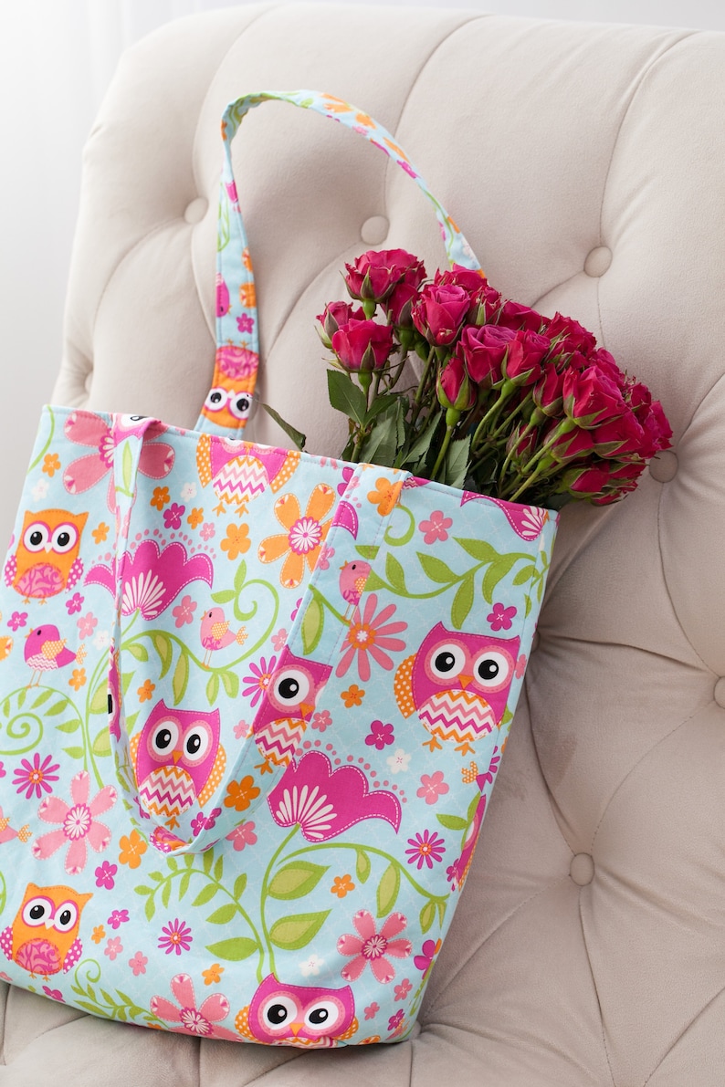 May include: A light blue tote bag with a colorful owl and floral pattern, filled with a bouquet of red roses. The bag's design includes pink, orange, and green accents, creating a cheerful and vibrant aesthetic.