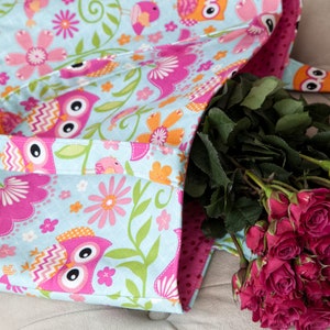 May include: A light blue tote bag with a colorful owl and floral pattern. The bag has a pink interior and a matching strap. A bouquet of red roses is placed inside the bag.