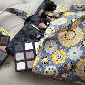 May include: A gray makeup bag with a floral pattern in yellow and white, next to a black quilted makeup brush holder and several eyeshadow palettes. The palettes contain various shades of purple, brown, and white eyeshadow.