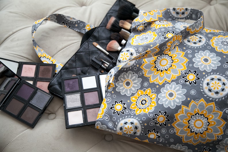 May include: A gray makeup bag with a floral pattern in yellow and white, next to a black quilted makeup brush holder and several eyeshadow palettes. The palettes contain various shades of purple, brown, and white eyeshadow.