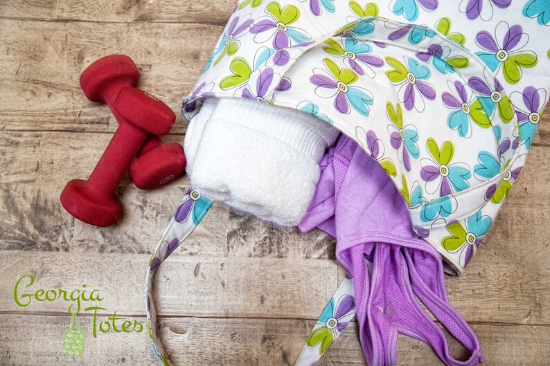 May include: A floral print tote bag with a white background and purple, green, and blue flowers. The bag is open, revealing a white towel and purple workout clothes. Two red dumbbells are next to the bag.